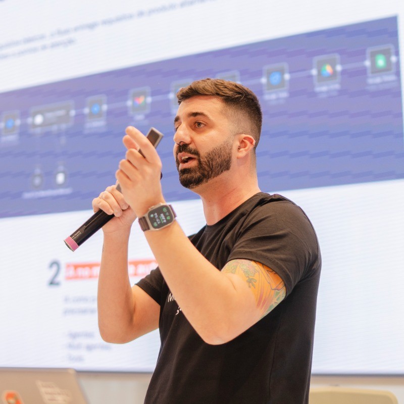 Kayo Ridolfi falando em uma palestra sobre Gen AI, microfone na mão e slide ao fundo.